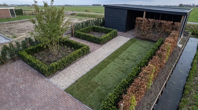 Luchtopname van een pas aangelegde tuin met bestrating, buxushagen, een grindpad, verse graszoden en een zwarte houten schuur, aan de rand van een polderkavel.
